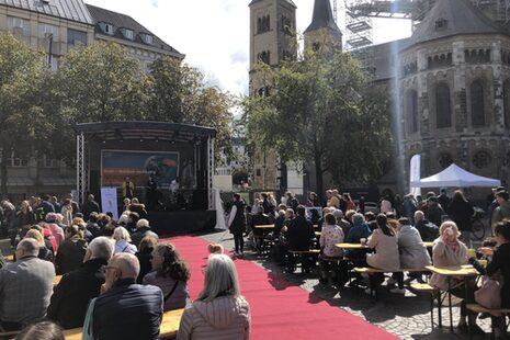 Publikum vor einer kleinen Open Air-Bühne, vor der als Laufsteg ein roter Teppich ausgerollt ist.