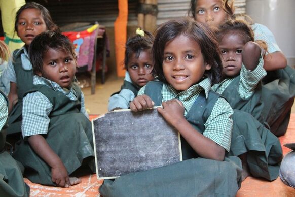 Mehrere Schulkinder in Indien schauen in die Kamera und zeigen eine Tafel