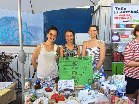 Drei junge Frauen stehen lächelnd hinter einem Informationsstand