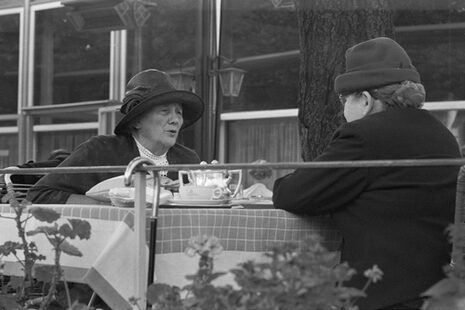 La foto en blanco y negro muestra a dos personas con sombreros y gorras. Están sentadas al aire libre en una mesa con tazas.