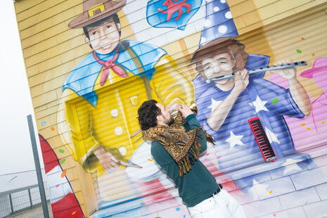 A man with a small trumpet in front of a mural with musicians