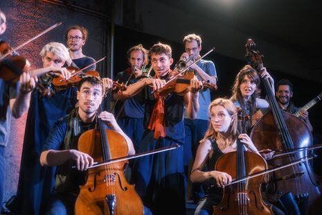 Violoncellisti e violinisti, sullo sfondo si intravedono persone con tromba