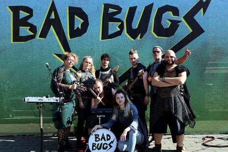 Punk kıyafetleri giymiş 8 kişi kameraya bakar. Biri grubun adını okuyor Bad Bugs