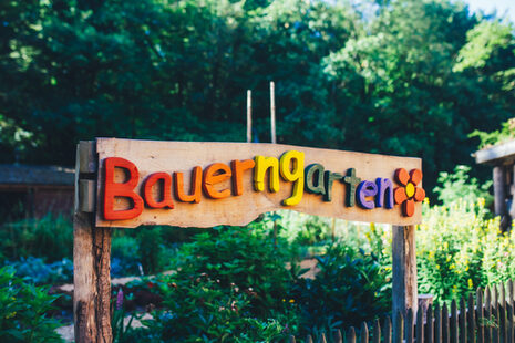 Das Foto zeigt ein Holzschild, auf dem in bunten Buchstaben „Bauerngarten“ steht. Dahinter ist ein Garten zu sehen.