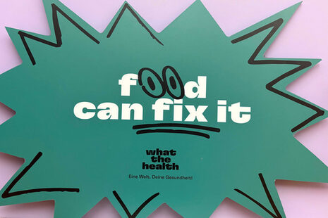 Das Foto zeigt ein Plakat mit einem stilisierten comicartigen Ausruf mit der Schrift: "food can fix it" in weißer Schrift, wobei die "os" von "food" als Augen dargestellt sind. Darunter steht in schwarzer Schrift: what the health. Eine Welt. Deine Gesundheit.