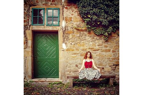 Das Foto zeigt ein altes Steinhaus, das mit Efeu bewachsen ist. Es hat eine grüne Tür und zwei grüne Sprossenfenster darüber. Auf einer Bank vor dem Haus sitzt eine Frau in Meditationshaltung.